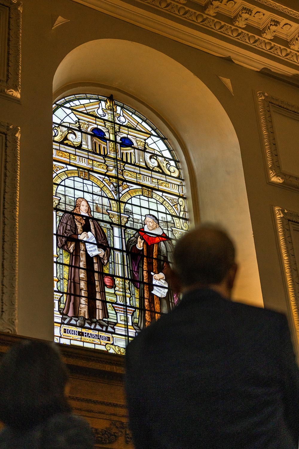 A man looks up at a stained glass window depicting John ŷպƵ and another scholar.